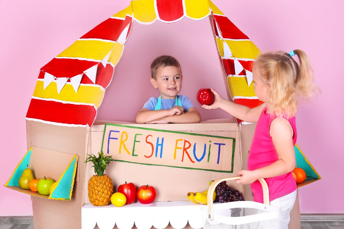 Little Children Playing with Cardboard Fruit Stall Against Pink Wall